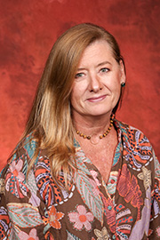 A smiling woman with blonde hair and glasses wearing a dark blue top poses against a reddish-brown background.